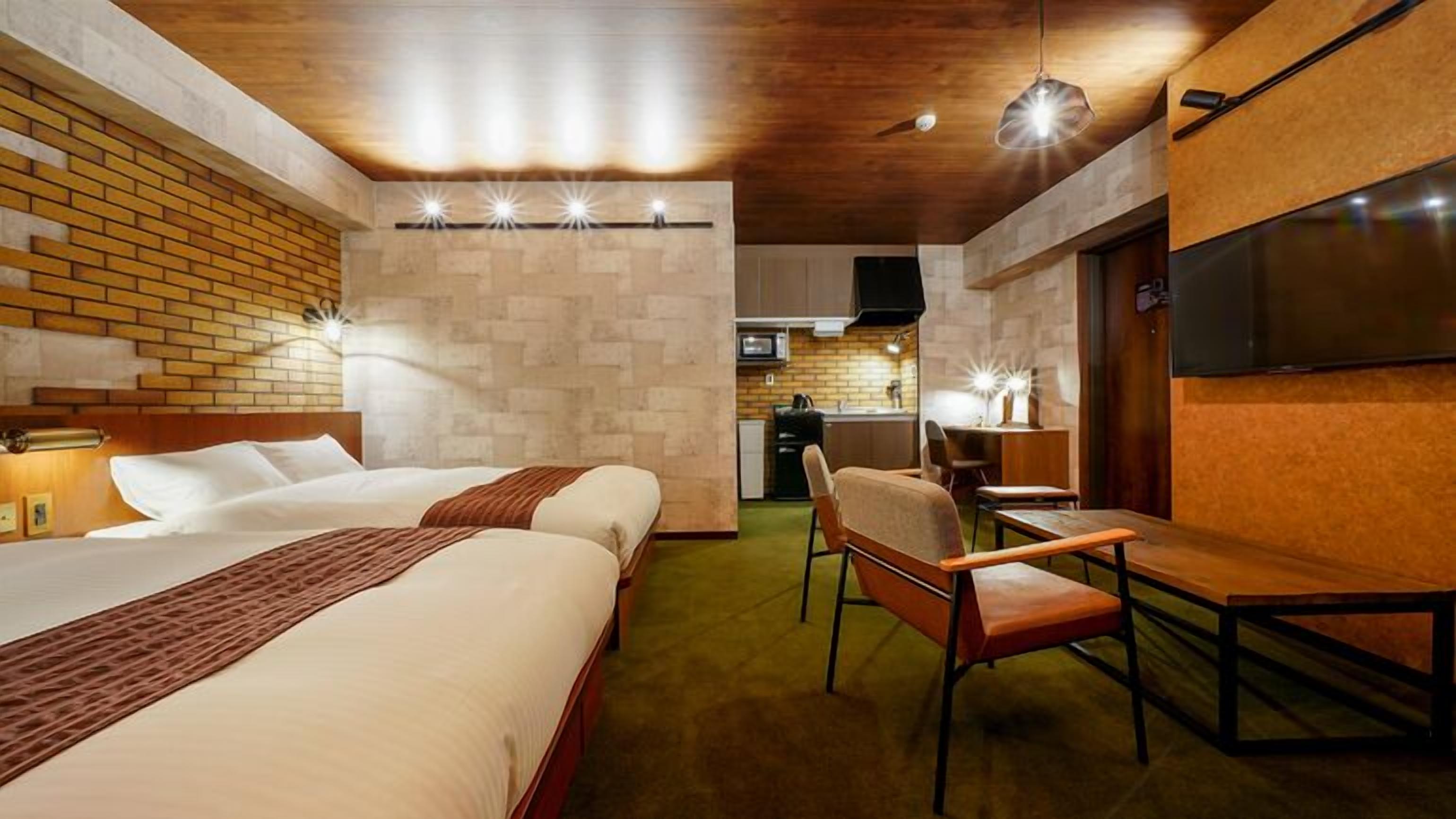 A hotel room with two beds, a desk, a chair, a TV, and a kitchenette.