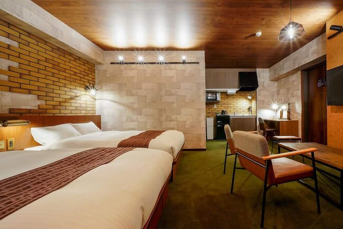 A hotel room with two beds, a desk, a chair, a TV, and a kitchenette.