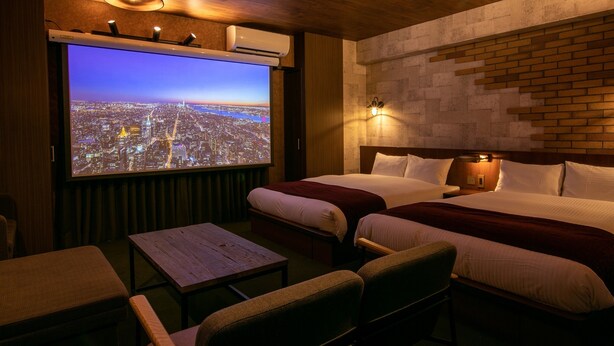 A hotel room with two beds, a large TV screen displaying a cityscape, a wooden ceiling, and a brick wall. - RESI STAY BRiX (Kyoto)