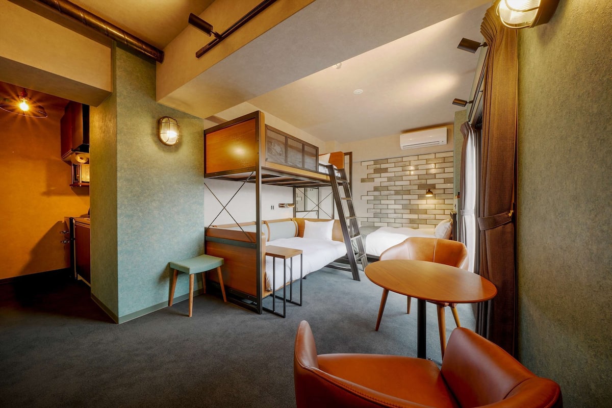 A compact hotel room with a bunk bed, a small desk, a chair, and a round table.