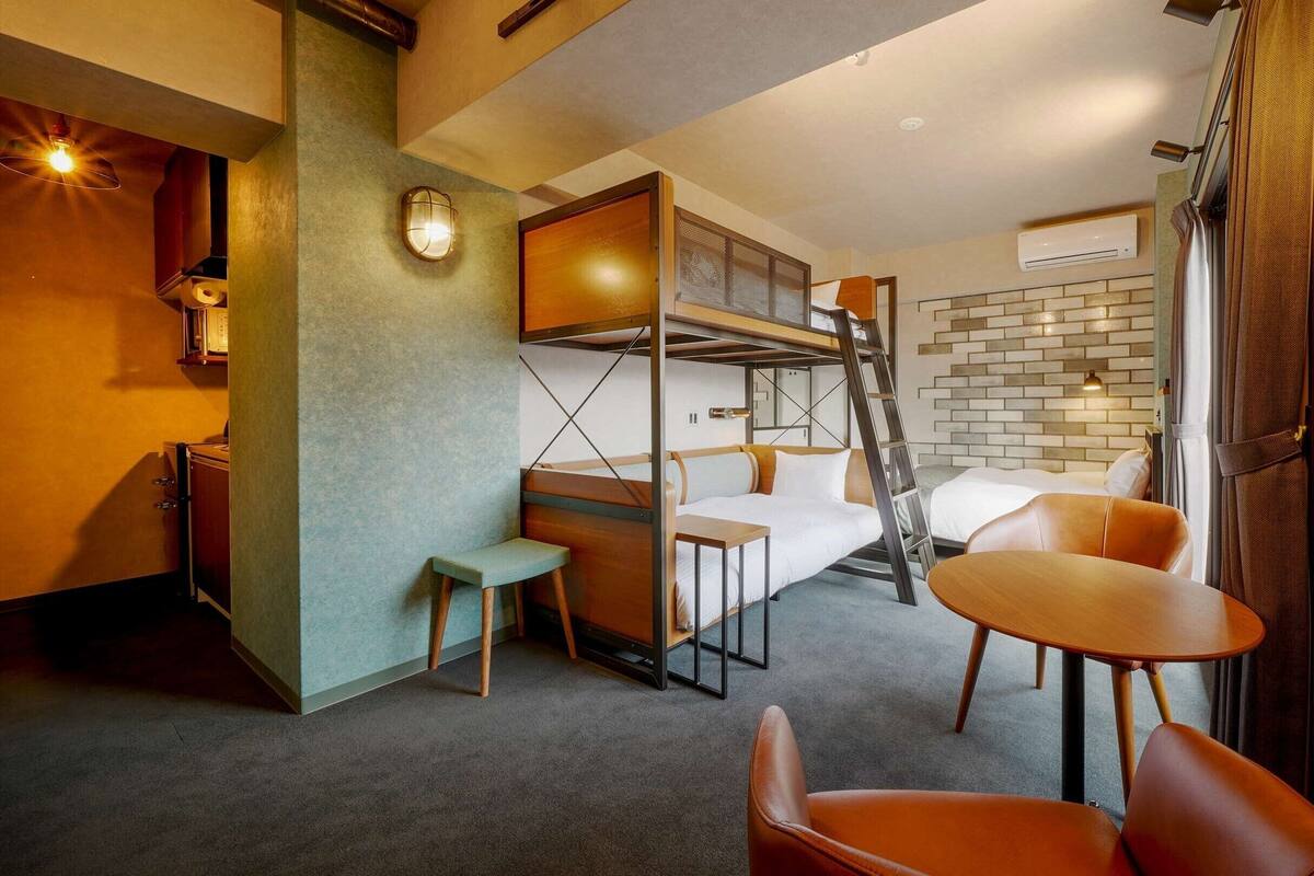 A compact hotel room with a bunk bed, a small table, a chair, and a sofa.