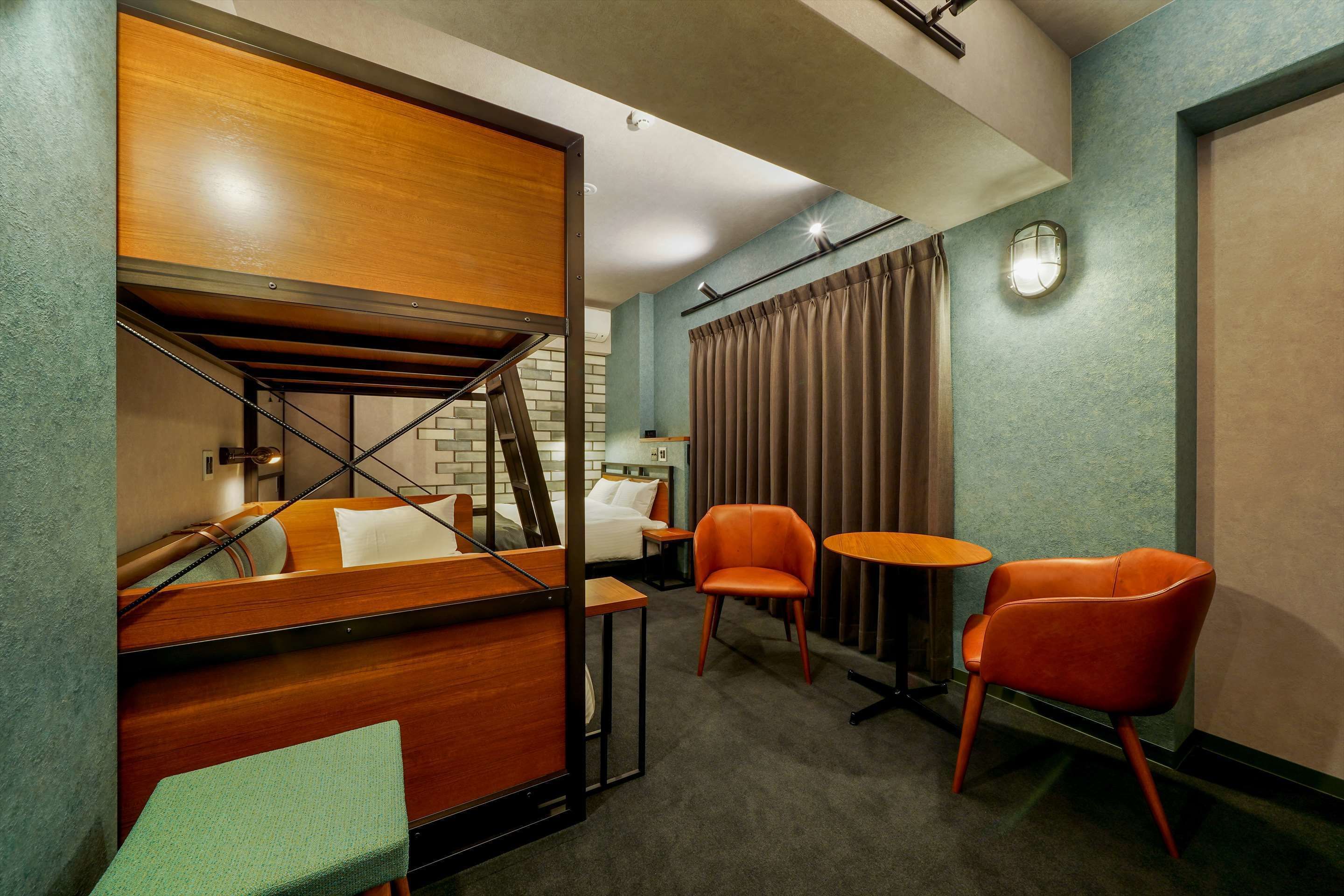 A hotel room with a wooden bunk bed, a desk, two chairs, a small table, and a curtain.