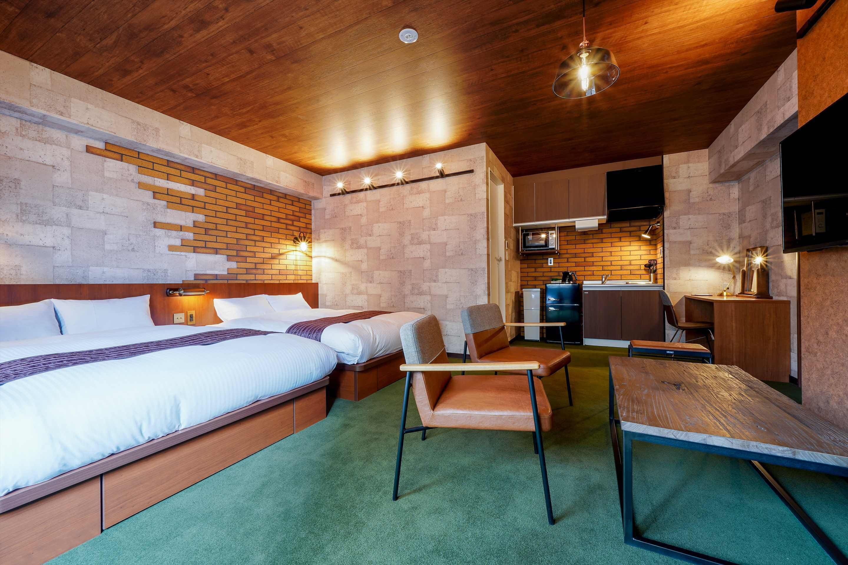 Quarto de hotel moderno com teto de madeira, parede com padrão de tijolos, cama grande, escrivaninha e cozinha compacta.