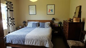 A neatly made bed with patterned bedding, a wooden dresser, a nightstand with a lamp, and two framed artworks on the wall.