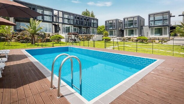 Un área de piscina moderna con terraza de madera, piscina con agua azul cristalina y edificios contemporáneos al fondo.