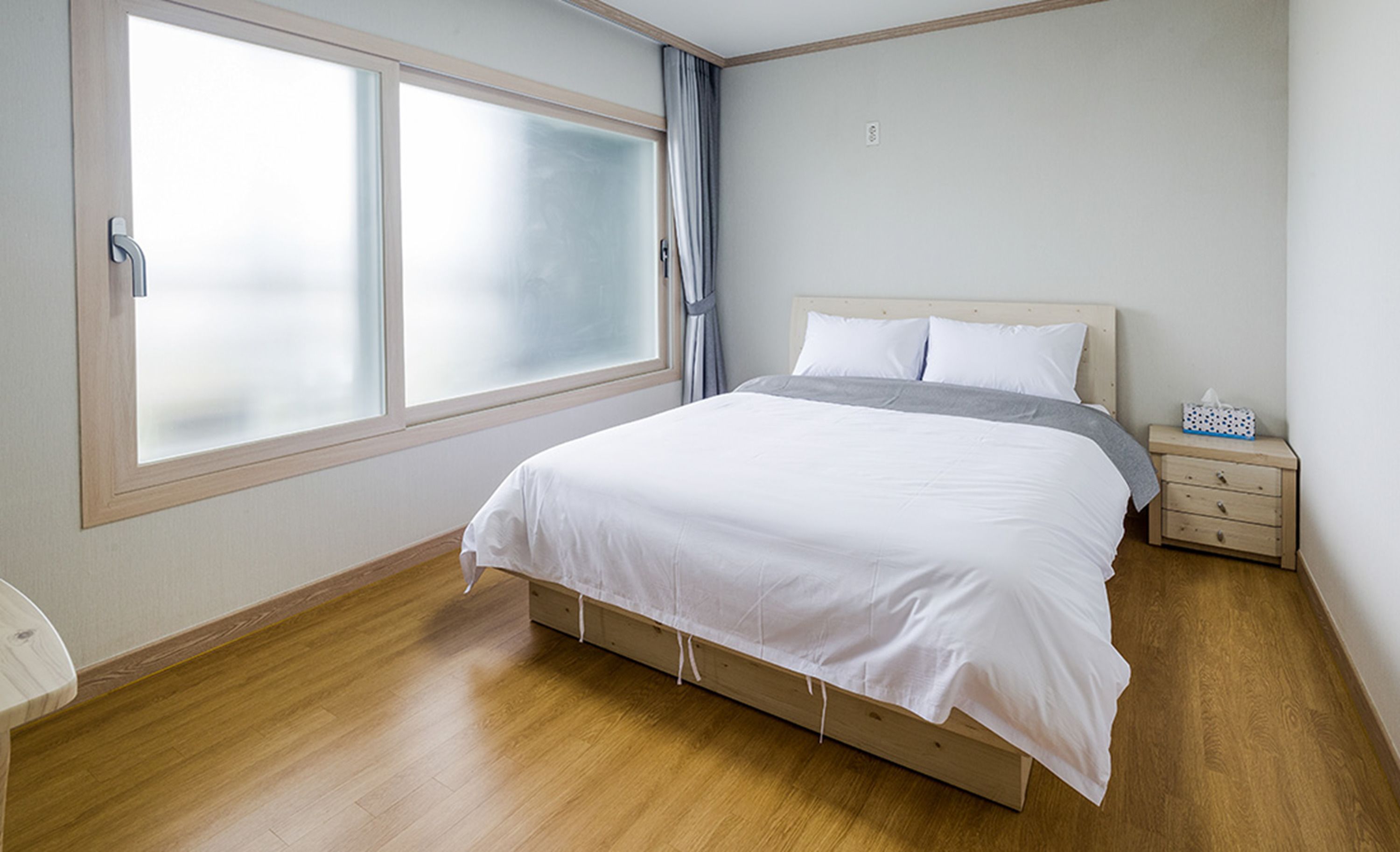A bedroom with a large window, a bed with white bedding, a wooden nightstand, and a small bedside table with a tissue box.
