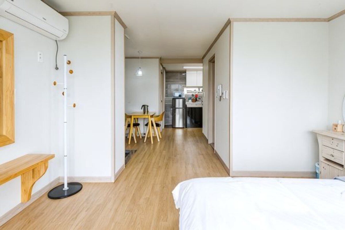 A hotel room with a bed, a desk, a chair, and a kitchenette in the background.