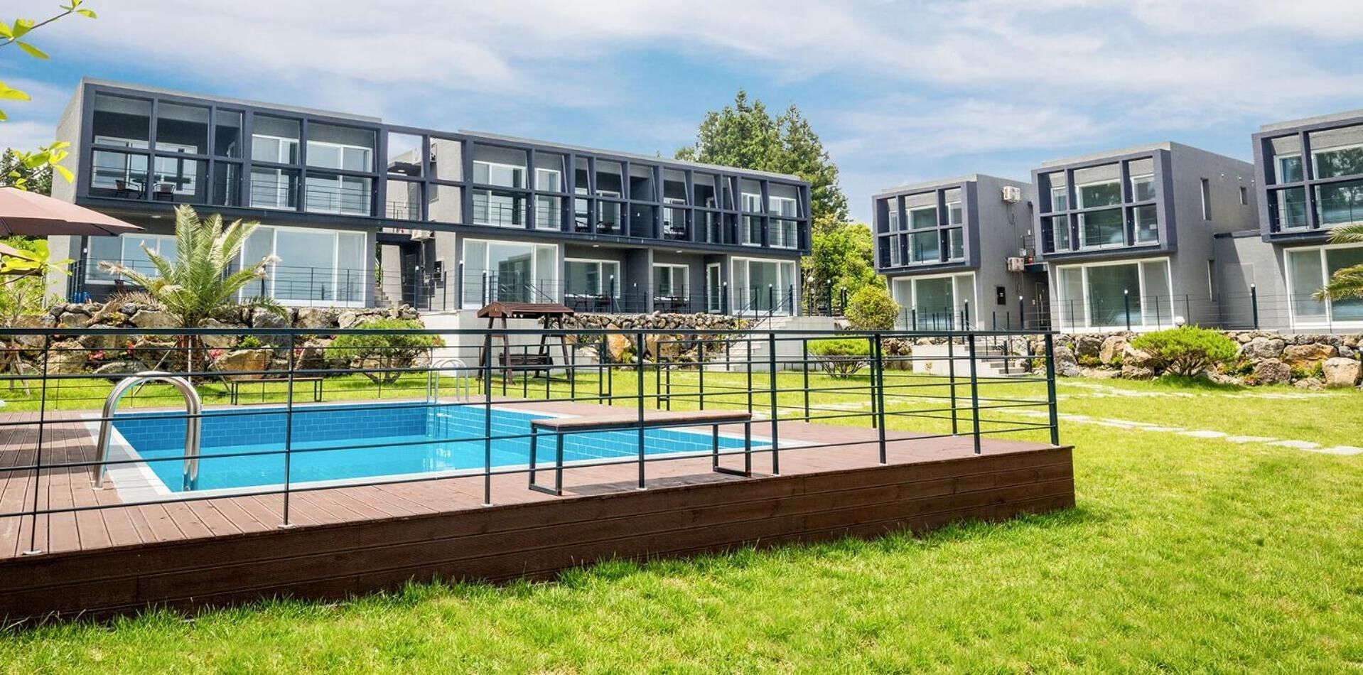 A modern residential building with a swimming pool, surrounded by a well-maintained lawn and a metal fence.