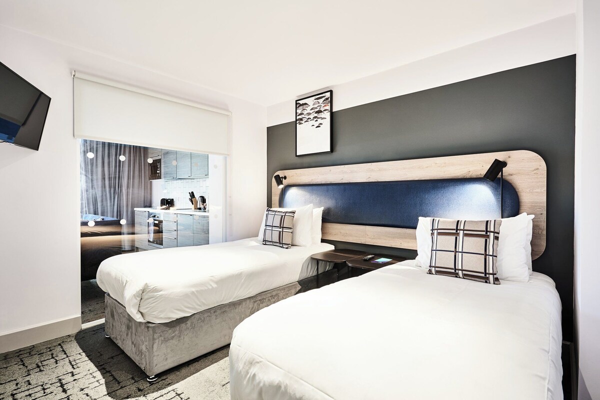 A hotel room with two beds, a TV, a small kitchen area, and a view of the outside.