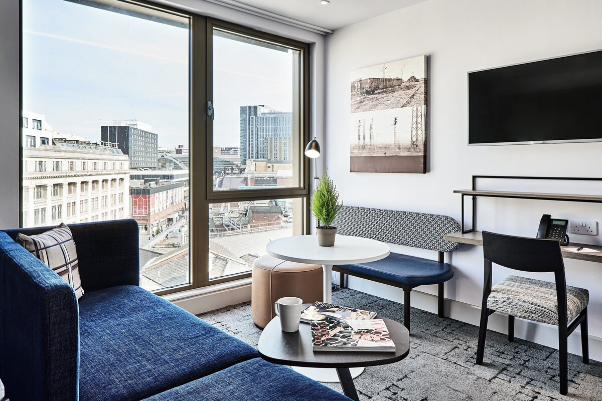 A modern hotel room with a large window offering a city view, a sofa, a small table with a magazine, a chair, and a desk with a phone.