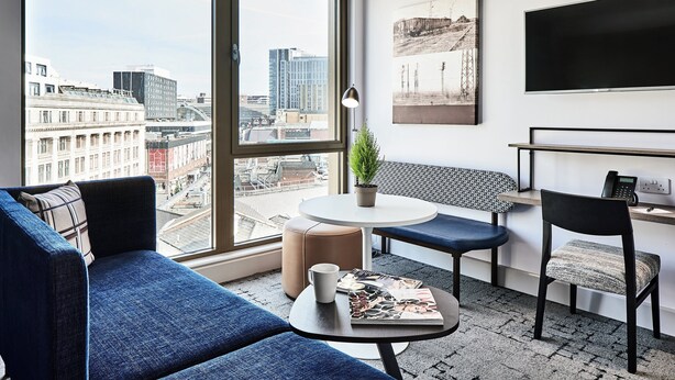 A modern hotel room with a large window offering a city view, a sofa, a small table with a magazine, a chair, and a desk with a phone.