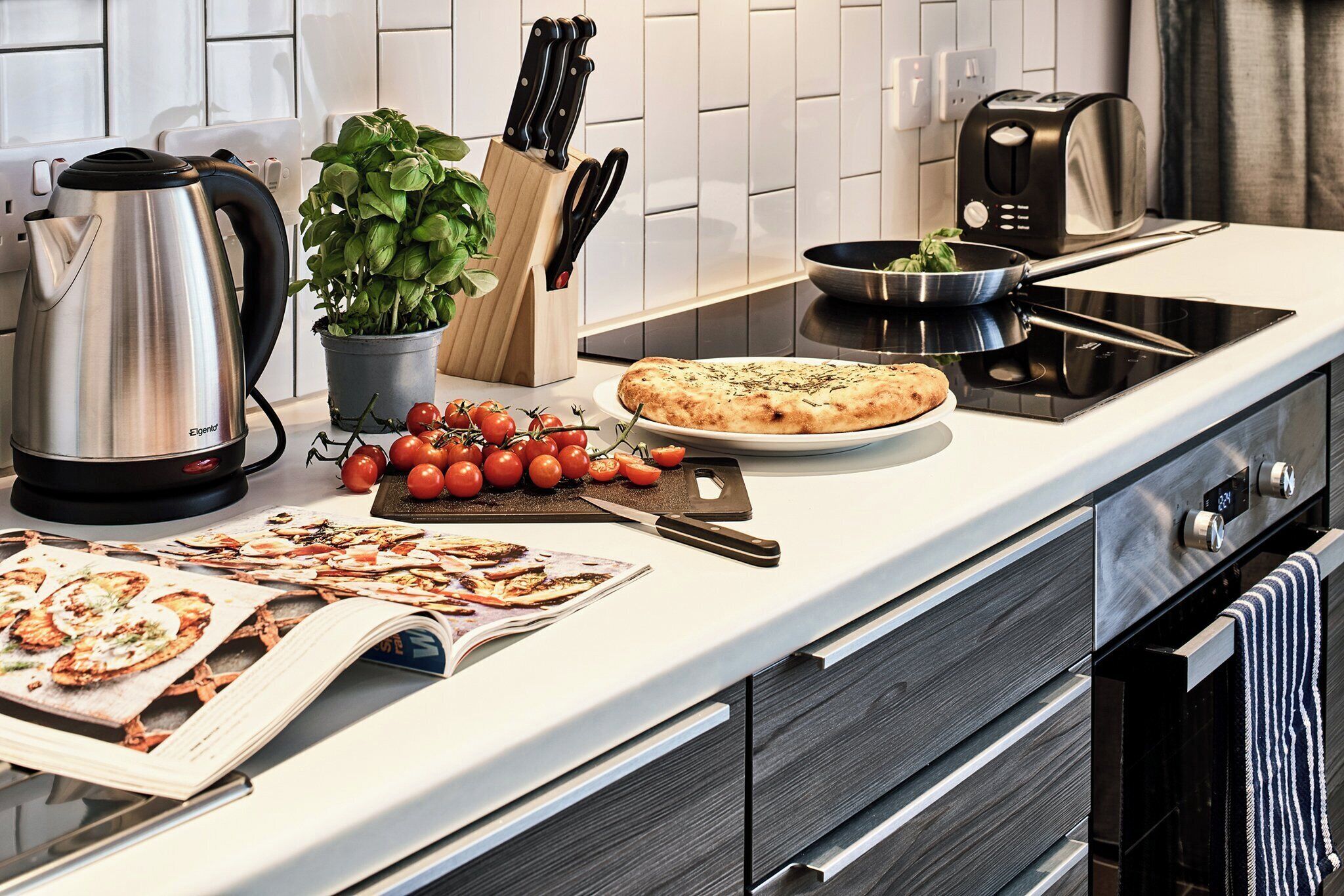 A modern kitchen with a stainless steel kettle, a potted basil plant, a cutting board with tomatoes, a pizza on a plate, and a magazine on the counter.