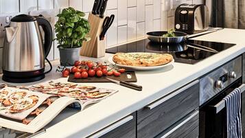A modern kitchen with a stainless steel kettle, a potted basil plant, a cutting board with tomatoes, a pizza on a plate, and a magazine on the counter.