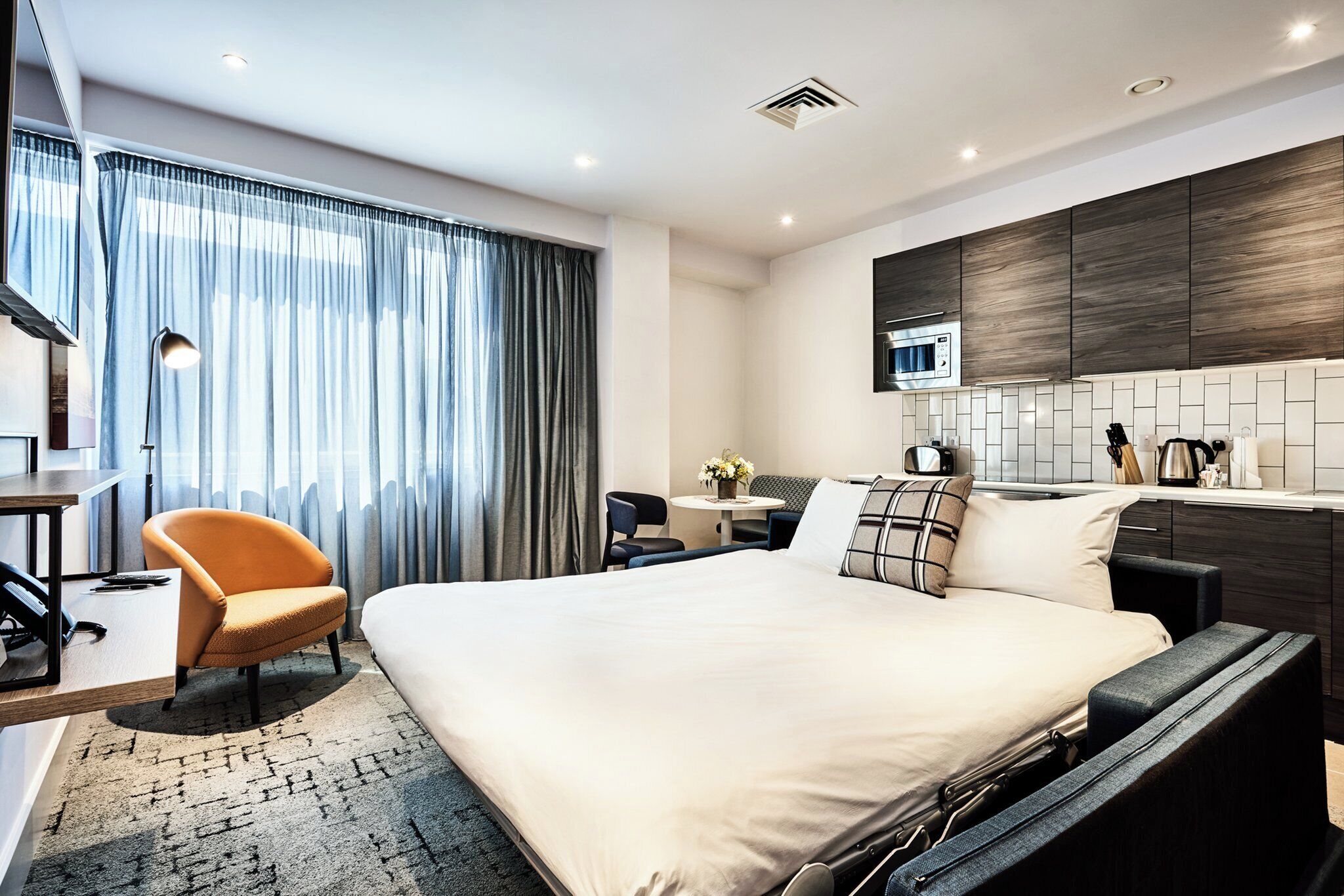 Een moderne hotelkamer met een groot bed, een bureau, een stoel, een bijzettafeltje met een bloemenvaas en een kitchenette met een magnetron.