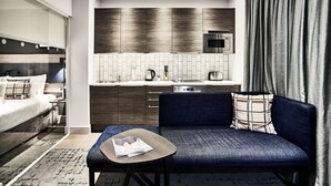Ein modernes Hotelzimmer mit Sofa, einem kleinen Tisch, einem Bett, einer Küchenzeile und einem großen Fenster mit Vorhängen.