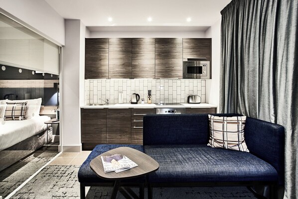 Una habitación de hotel moderna con sofá, una pequeña mesa, cama, kitchenette y un ventanal con cortinas.