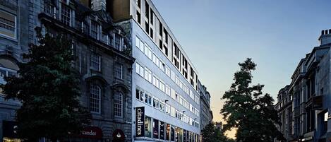 Un edificio histórico con fachada de vidrio moderna, una zona peatonal con árboles y comercios, incluyendo 'citadines'.
