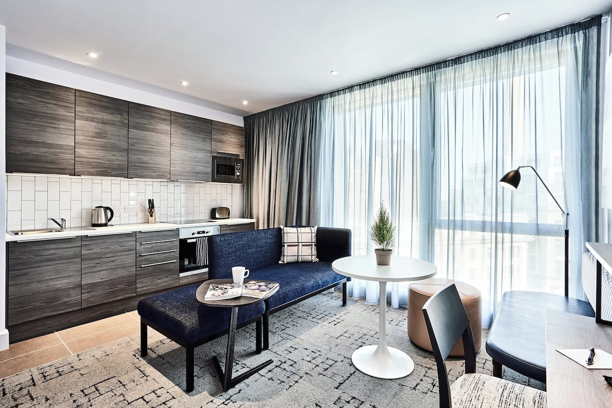 Una habitación de hotel moderna con kitchenette, un sofá, una mesa redonda y un amplio ventanal con cortinas.