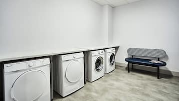 A laundry room with three washing machines, a bench, and a white wall.