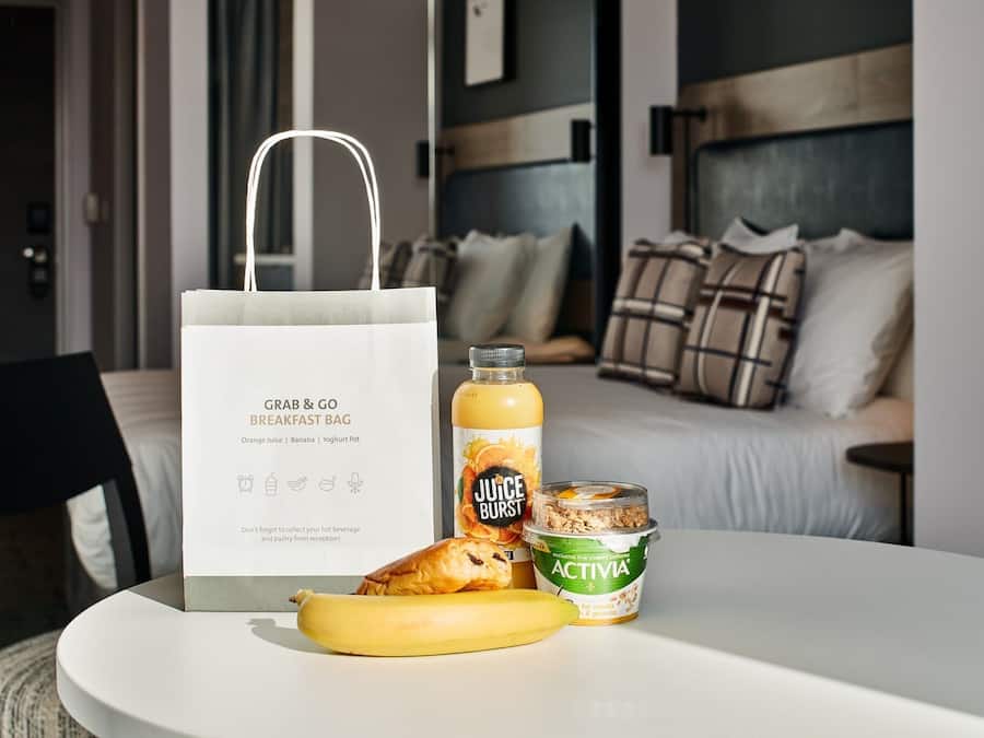 Un sac contenant le petit-déjeuner, une bouteille de jus d’orange, une banane et un pot de yaourt, posés sur une table dans une chambre d’hôtel.