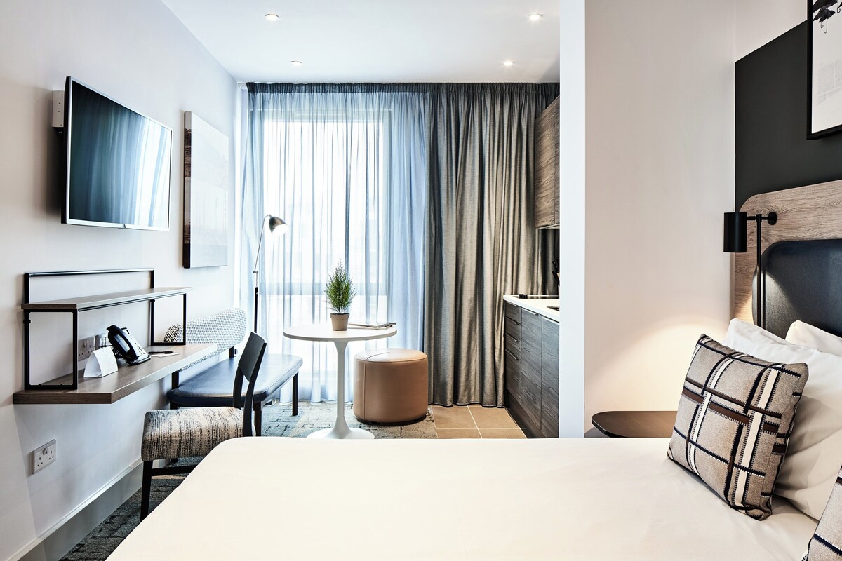A modern hotel room with a bed, a desk, a chair, a small table, and a TV.