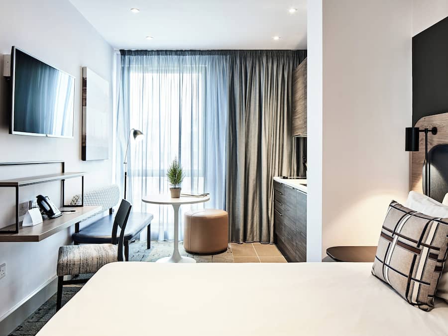 Quarto de hotel moderno com uma cama, uma escrivaninha, uma cadeira, uma mesinha e uma televisão.