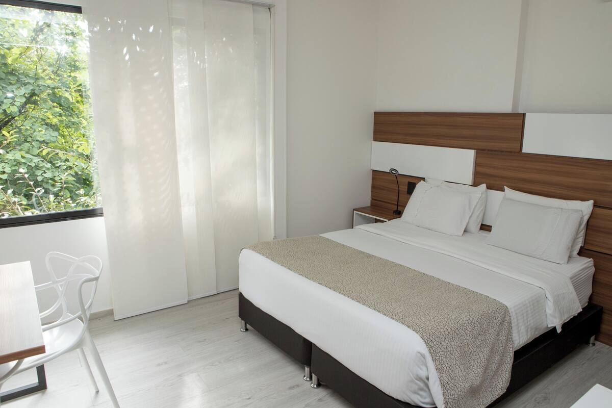 A modern hotel room with a large bed, a bedside table, a chair, and a window with curtains.