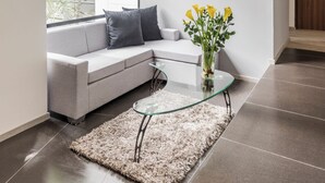 A modern living room with a grey sectional sofa, a glass coffee table, and a large window with a view of greenery. - Foresta Hotel (Medellín)