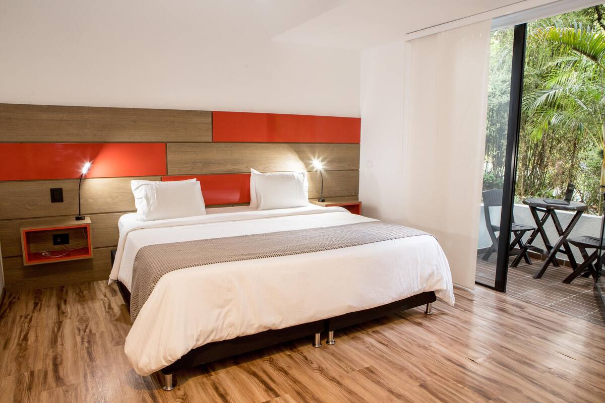 A modern hotel room with a large bed, wooden flooring, and a view of greenery through the glass door.