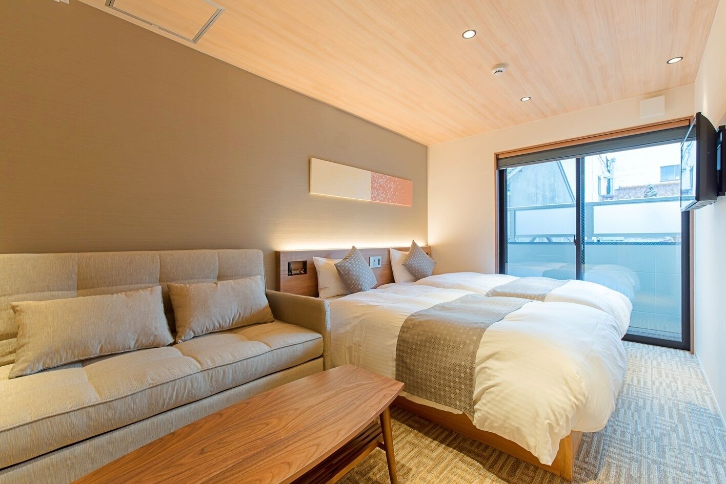 A modern hotel room with a sofa, a bed, a wooden table, and a large window.