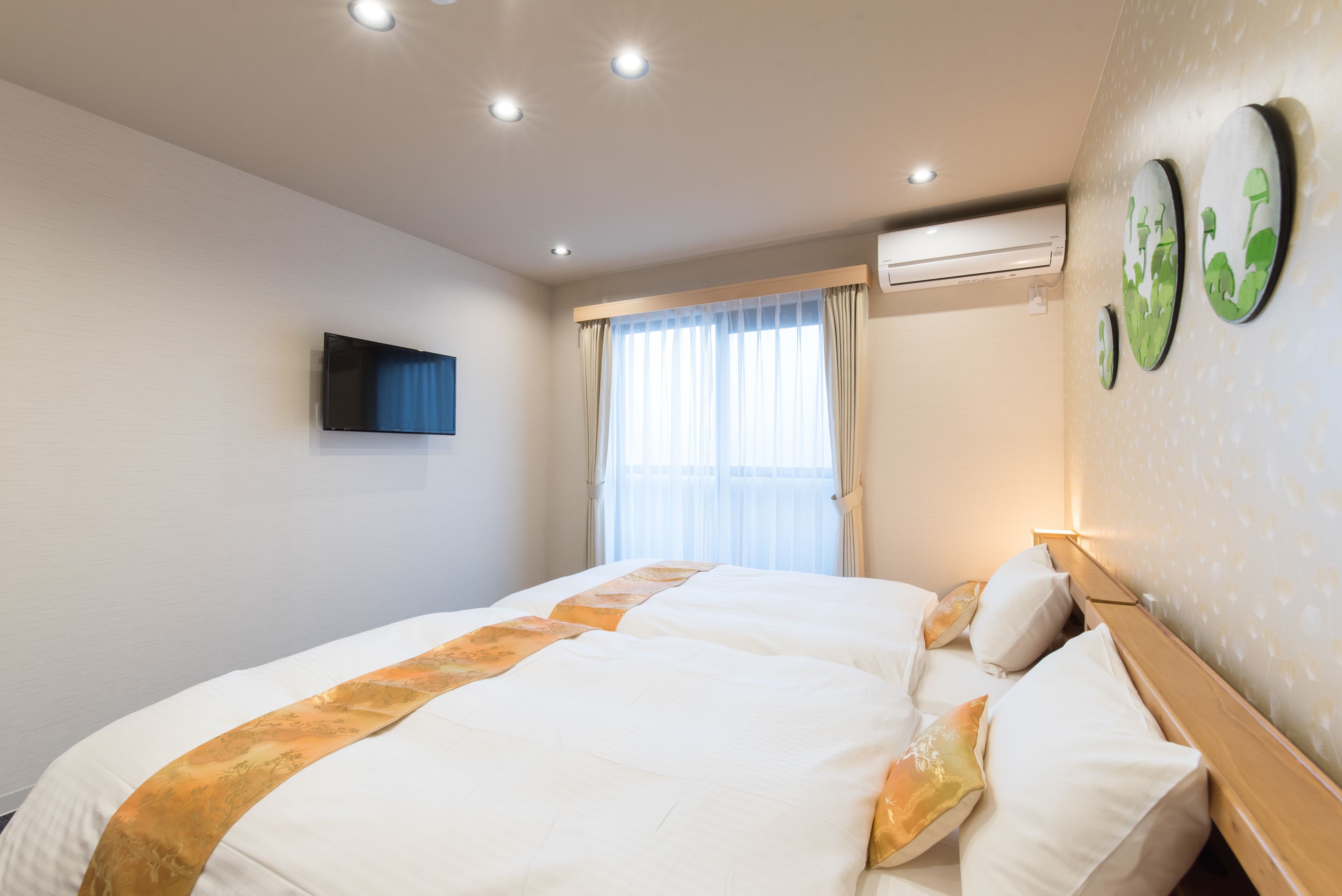 A hotel room with two beds, a flat-screen TV, an air conditioning unit, and a window with curtains.