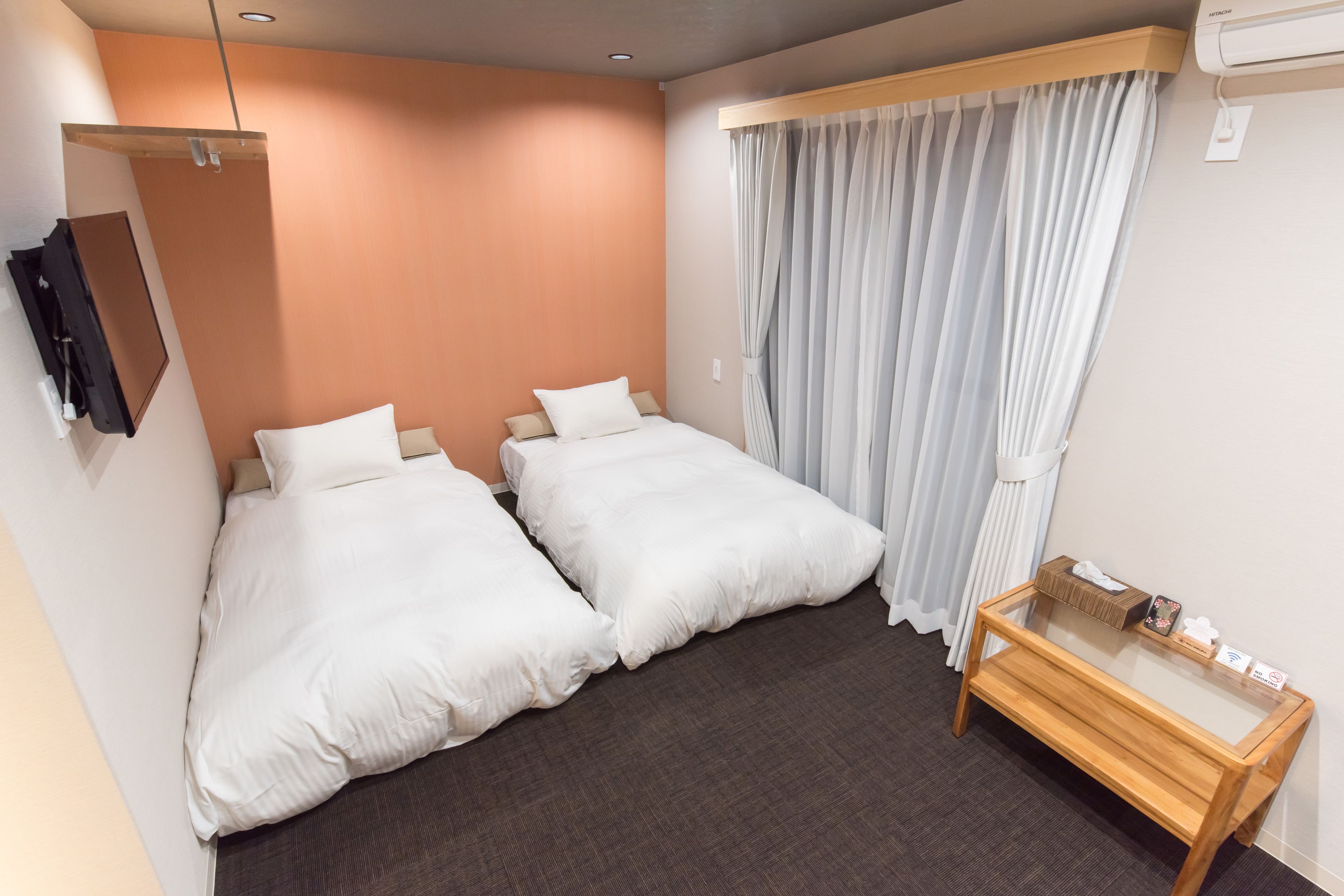 A hotel room with two beds, a small wooden table, a mirror, and a window with curtains.