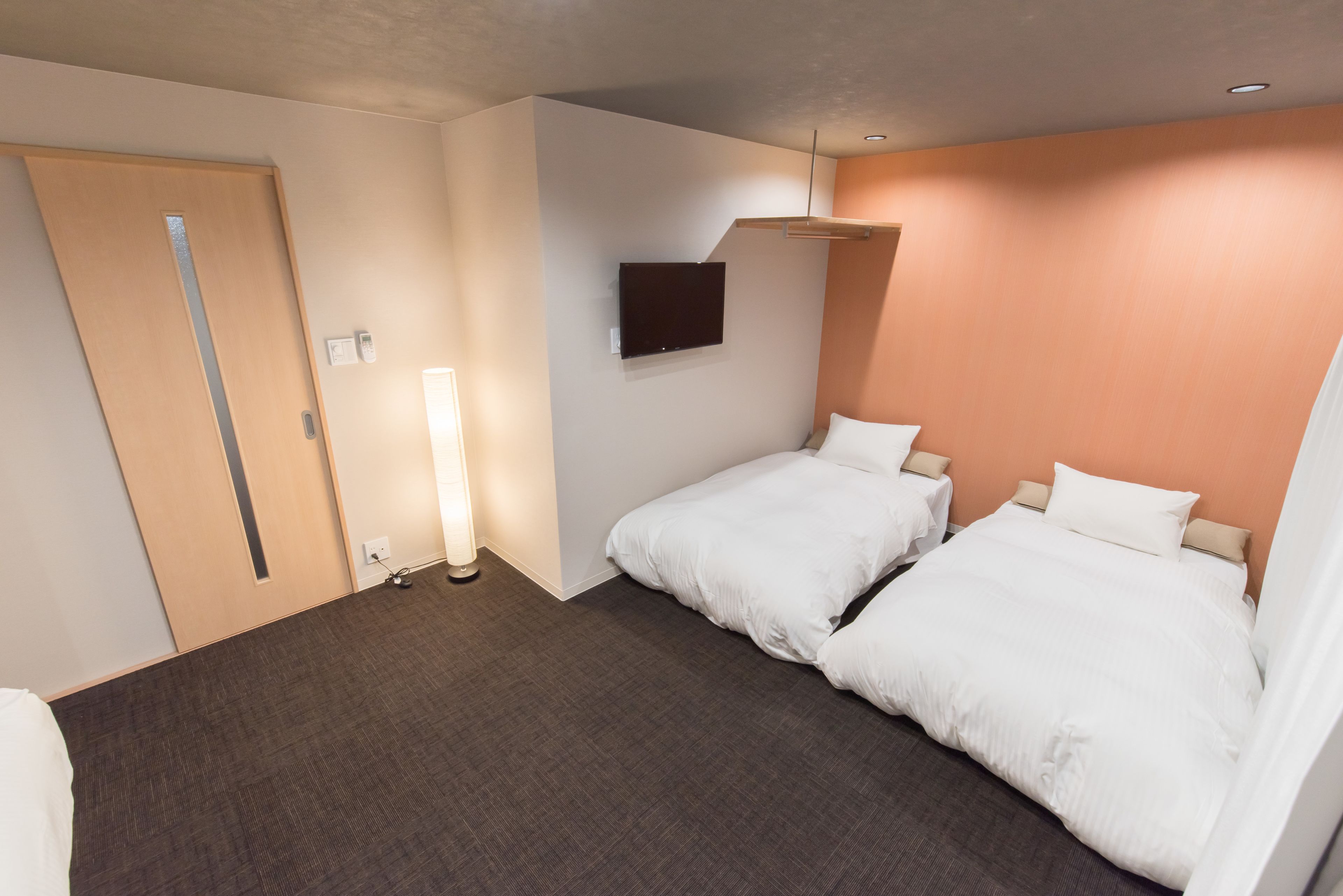 A hotel room with two beds, a flat-screen TV mounted on the wall, a door with a handle, and a bedside table with a lamp.
