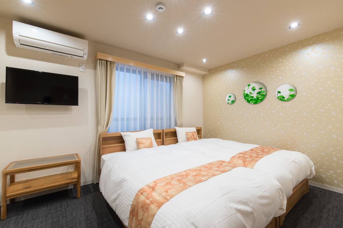 A hotel room with a large bed, a TV mounted on the wall, an air conditioning unit, and a window with curtains.