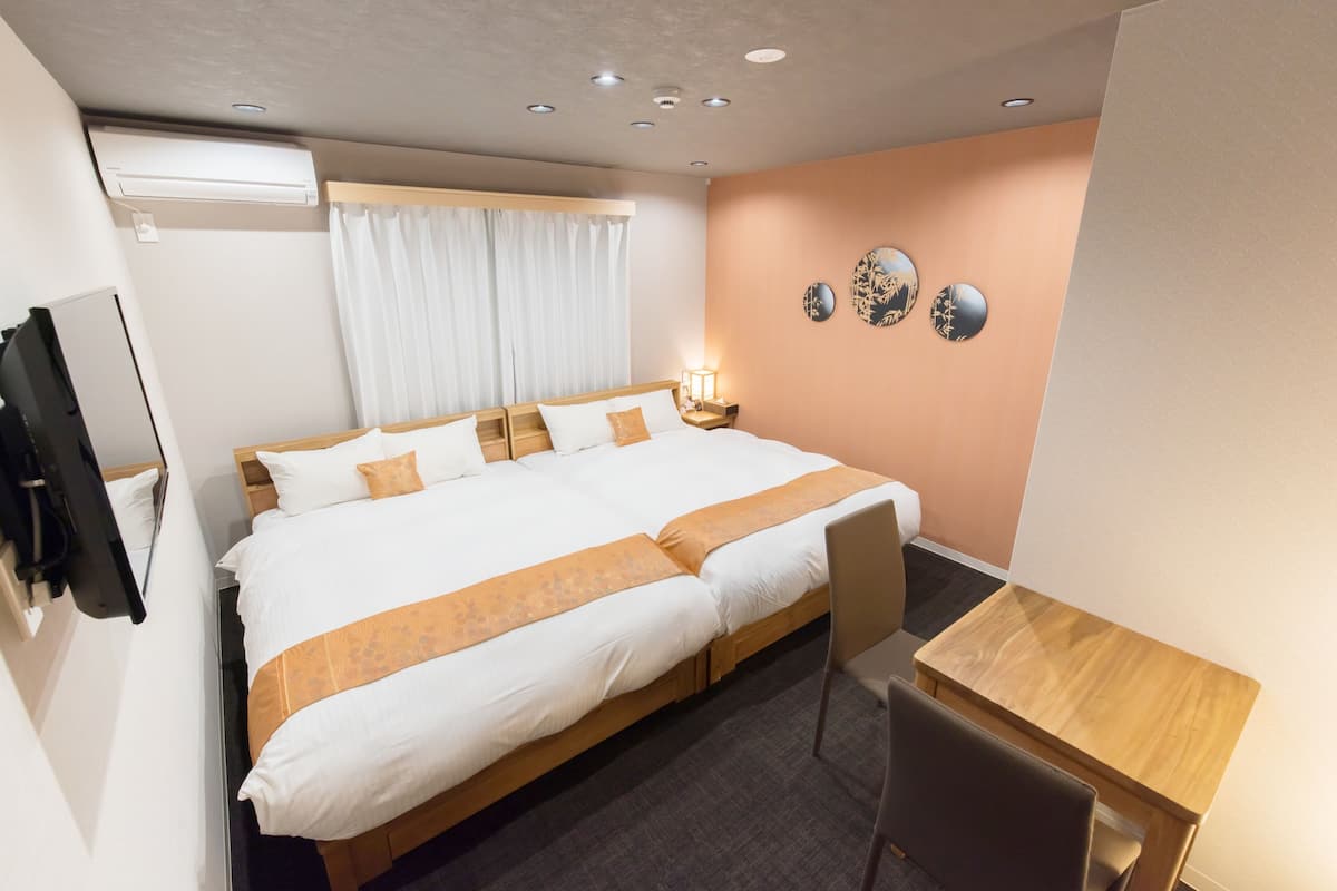 A hotel room with a bed, a desk, a chair, an air conditioner, a window with curtains, and wall decorations.