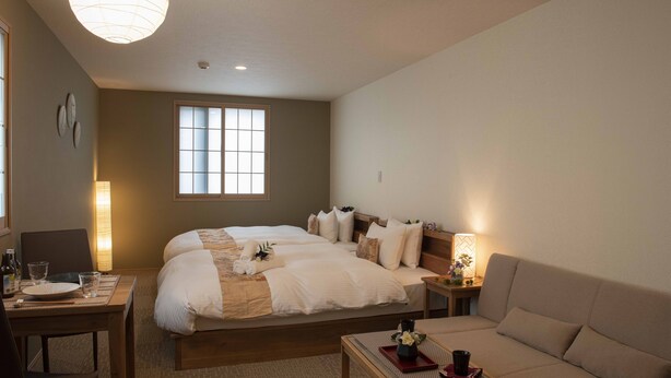 Habitación de hotel con una cama grande, una mesita de noche con lámpara, un sofá y una mesa de comedor dispuesta con vasos y platos.