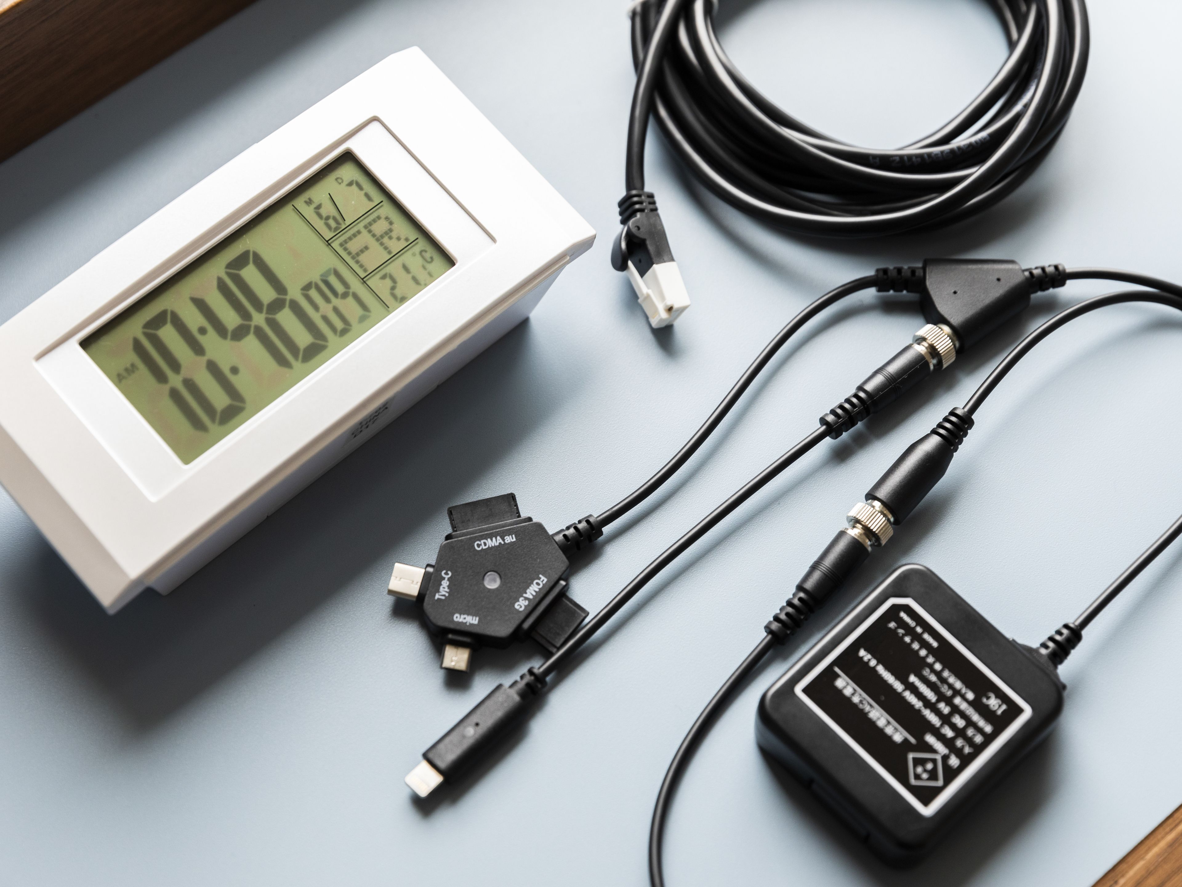 Un reloj digital que marca las 10:49, un cable USB y un adaptador de corriente sobre una superficie plana.
