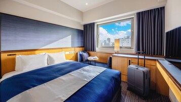 Habitación de hotel con una cama grande, un escritorio, una silla, una maleta y una ventana con vistas a la ciudad.