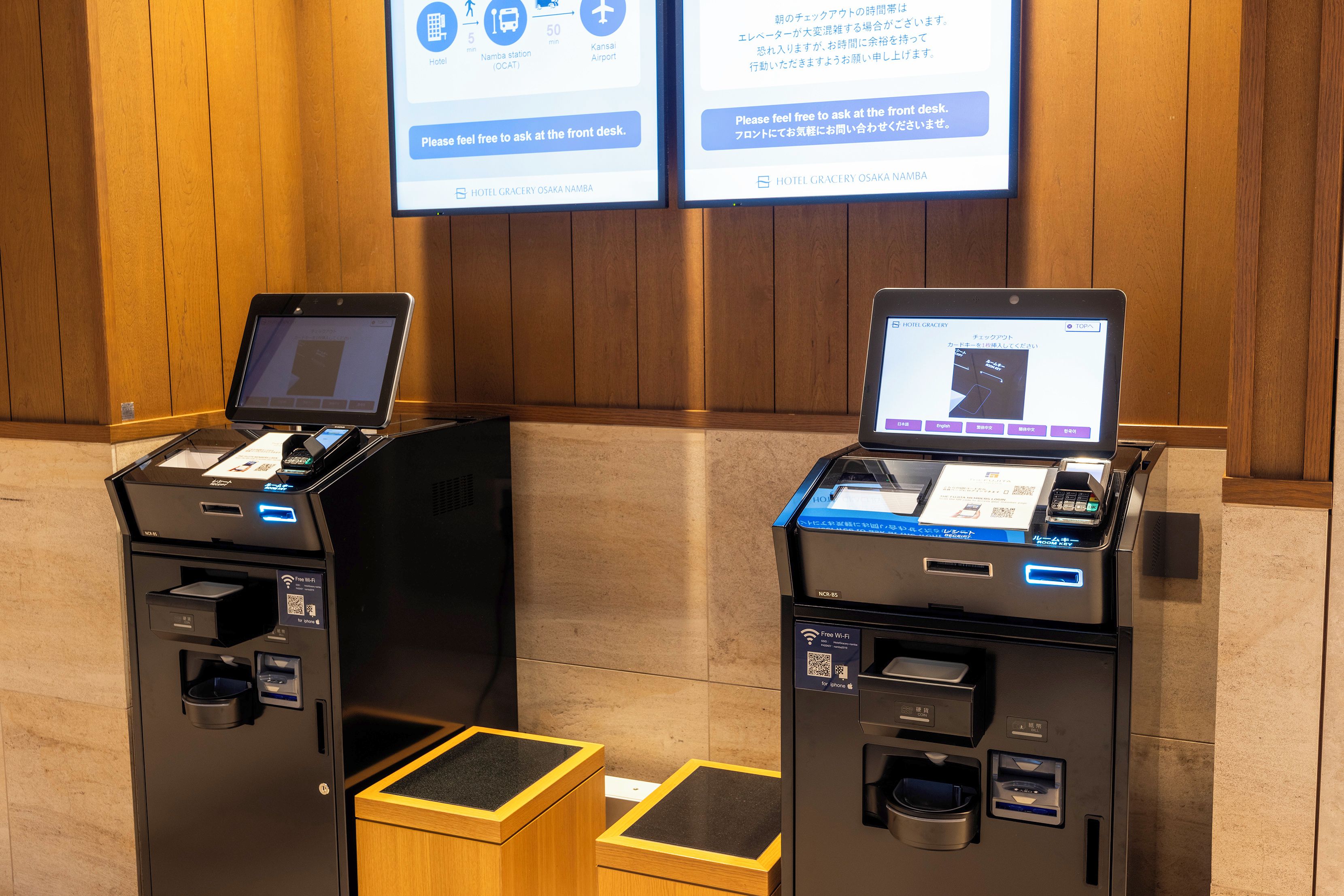 Two self-service kiosks with digital displays, a wooden floor, and a wooden wall.