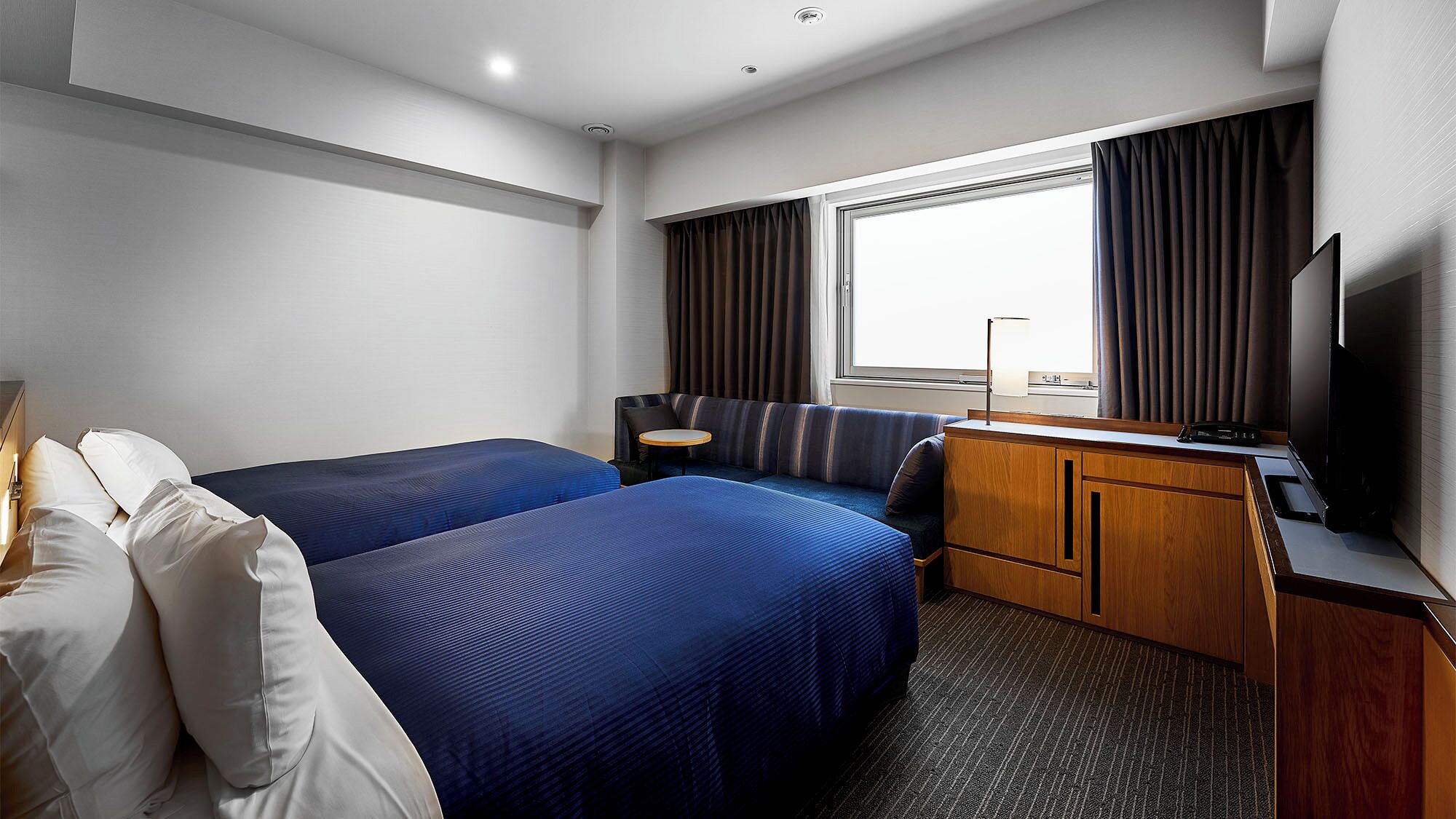 A hotel room with two beds, a desk, a TV, and a window with curtains.