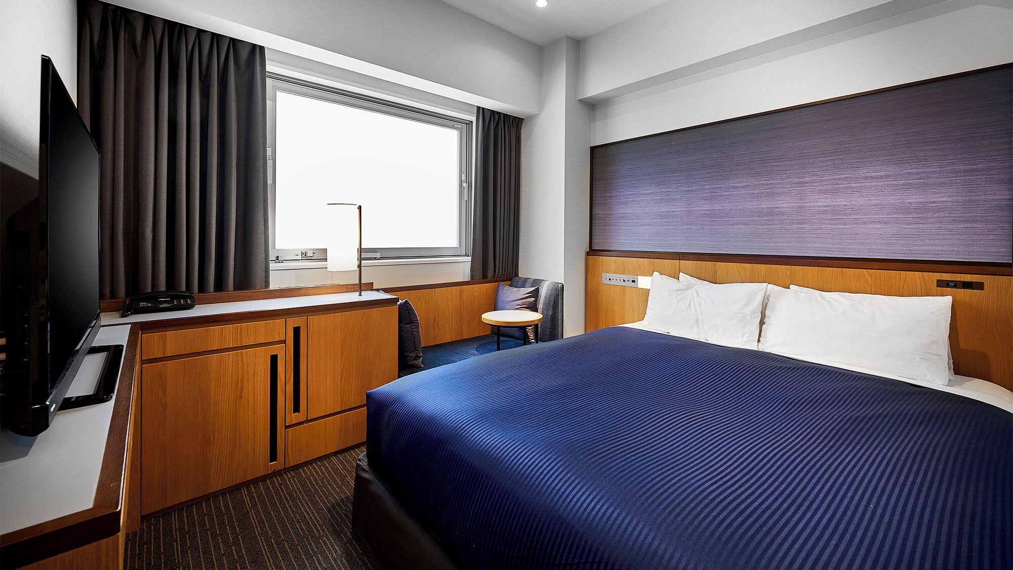 A hotel room with a bed, a desk, a chair, a TV, and a window with curtains.