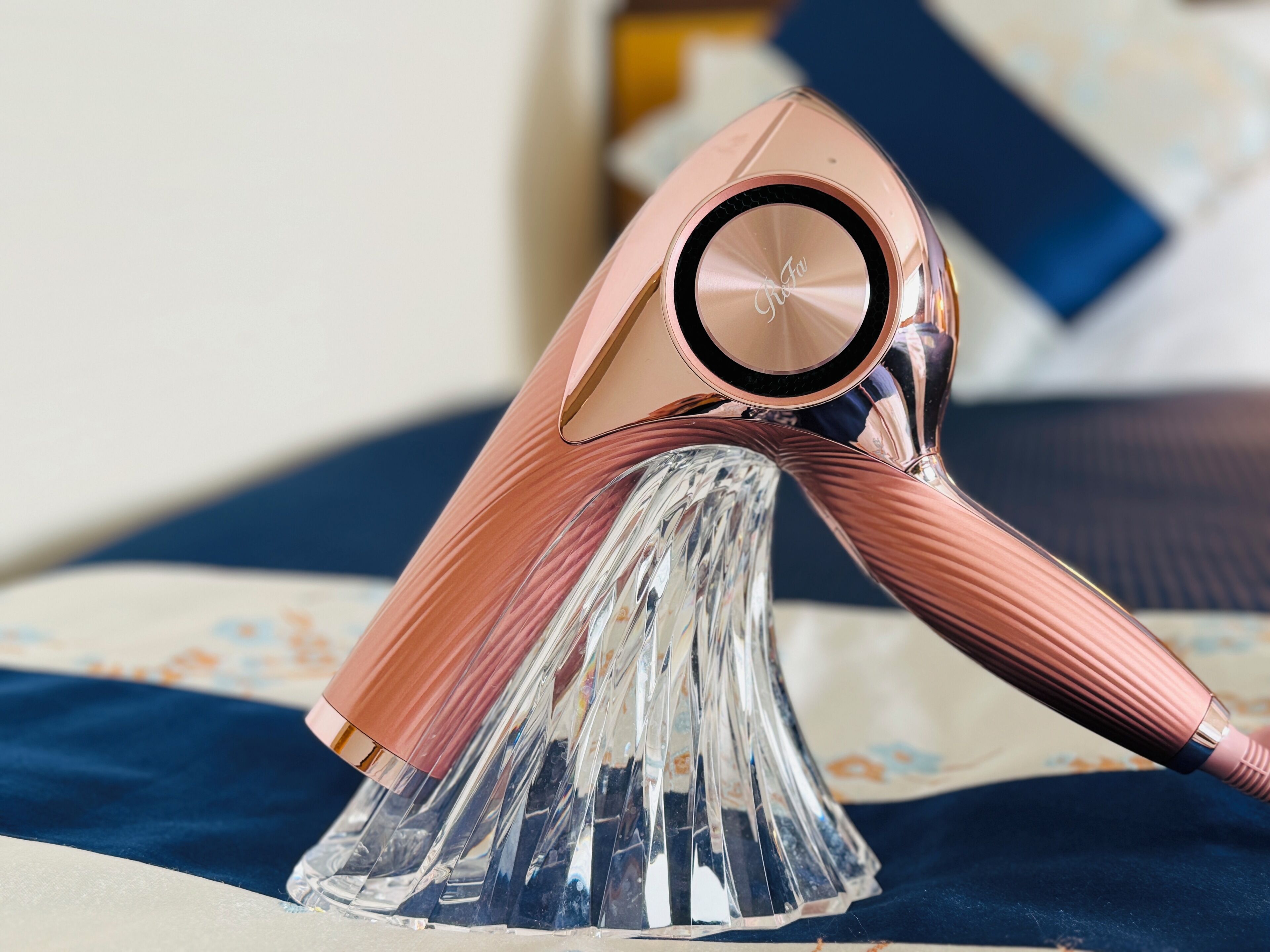 Un secador de pelo color rosa dorado con una base transparente y acanalada sobre una cama con ropa de cama estampada.
