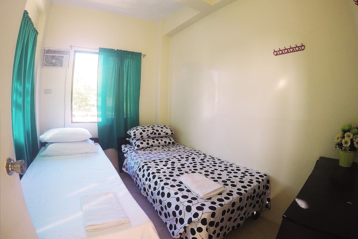 Habitación individual con una cama, una mesita de noche pequeña, una ventana con cortinas y un calentador fijado a la pared.