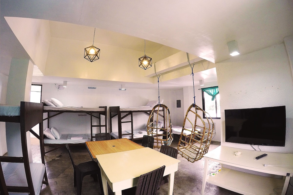 Una habitación con literas, una mesa de comedor con sillas, sillones colgantes de mimbre, un televisor y lámparas colgantes.