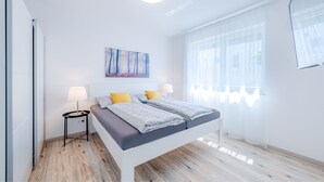 A modern bedroom with a bed, bedside tables, a painting, and a large window with sheer curtains.