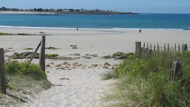 A sandy beach with a clear view of the ocean and a few people walking. There are rocks and some vegetation near the path leading to the beach. - Holiday house near the beaches (Penmarch)