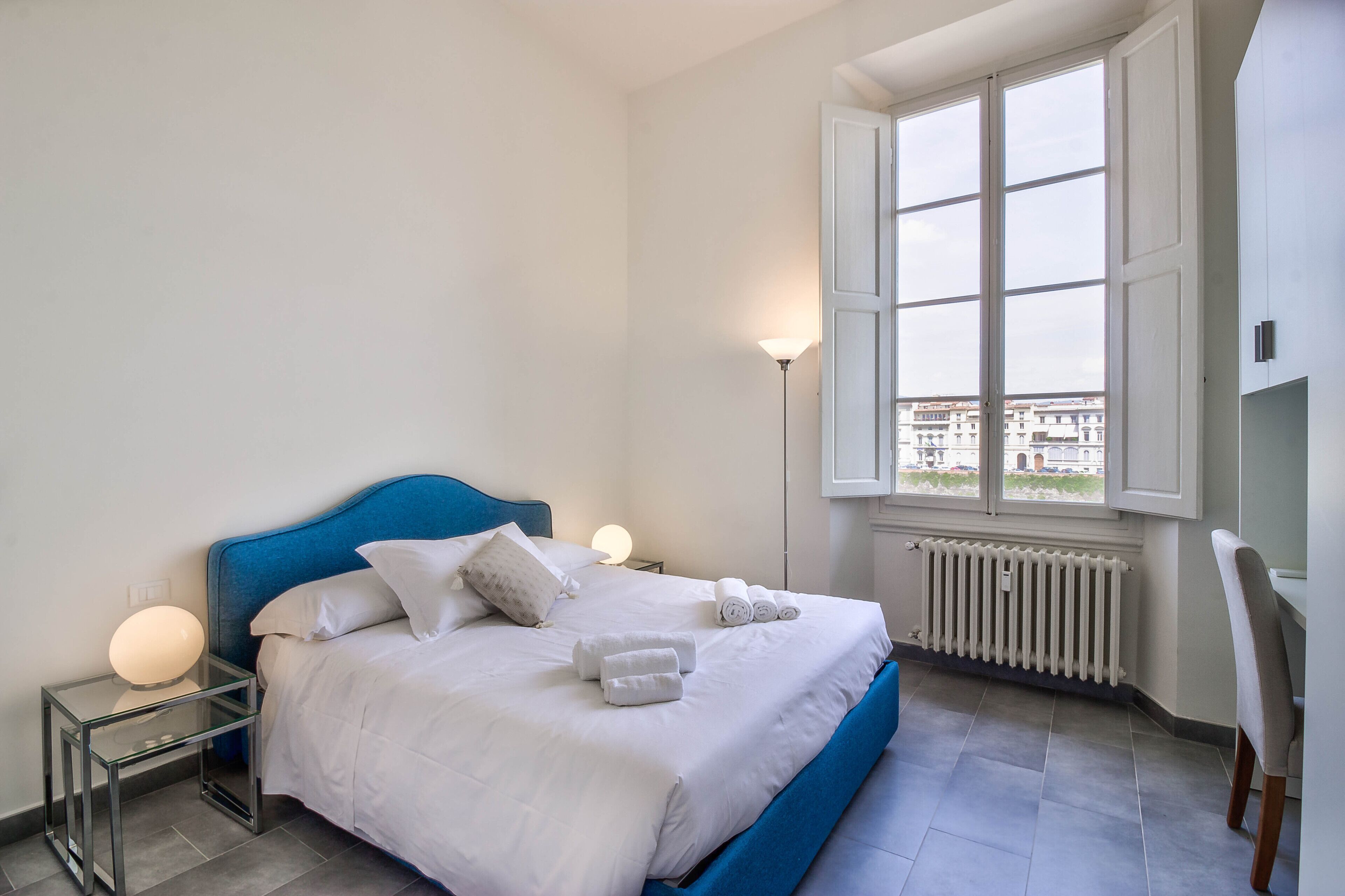 Een opgemaakt bed met witte lakens en opgerolde handdoeken, een blauw hoofdeinde, een bedlampje en een uitzicht op gebouwen door een raam.