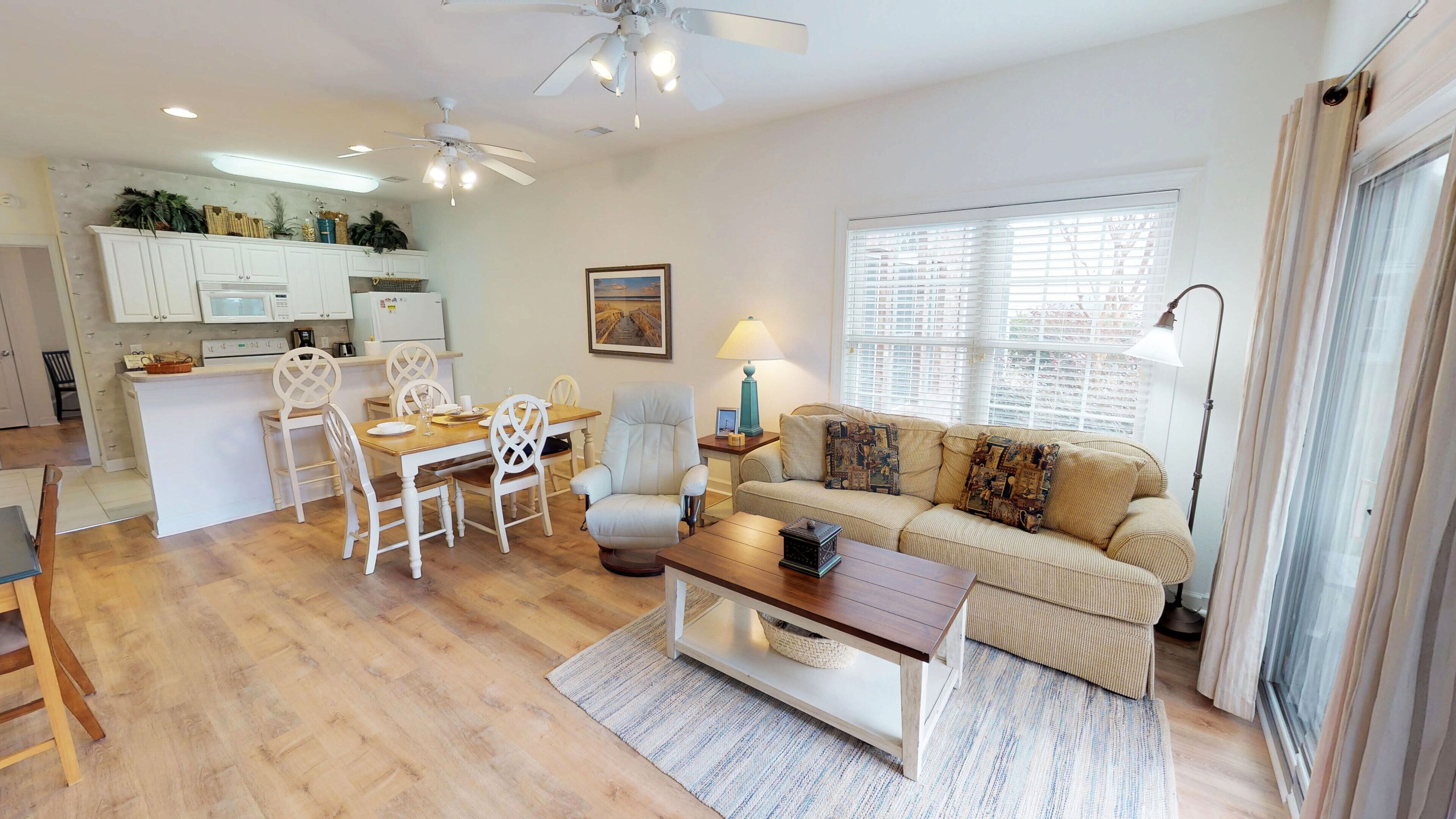 A modern living room with a wooden dining table, white chairs, a beige sofa, and a coffee table. There is a kitchen area with white cabinets and a microwave.