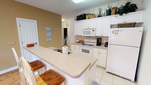 A kitchen with white cabinets, a microwave, a refrigerator, and a wooden dining table with two chairs. - CH3702, Tee Off in Comfort, Perfect Golf Retreat (Sunset Beach)