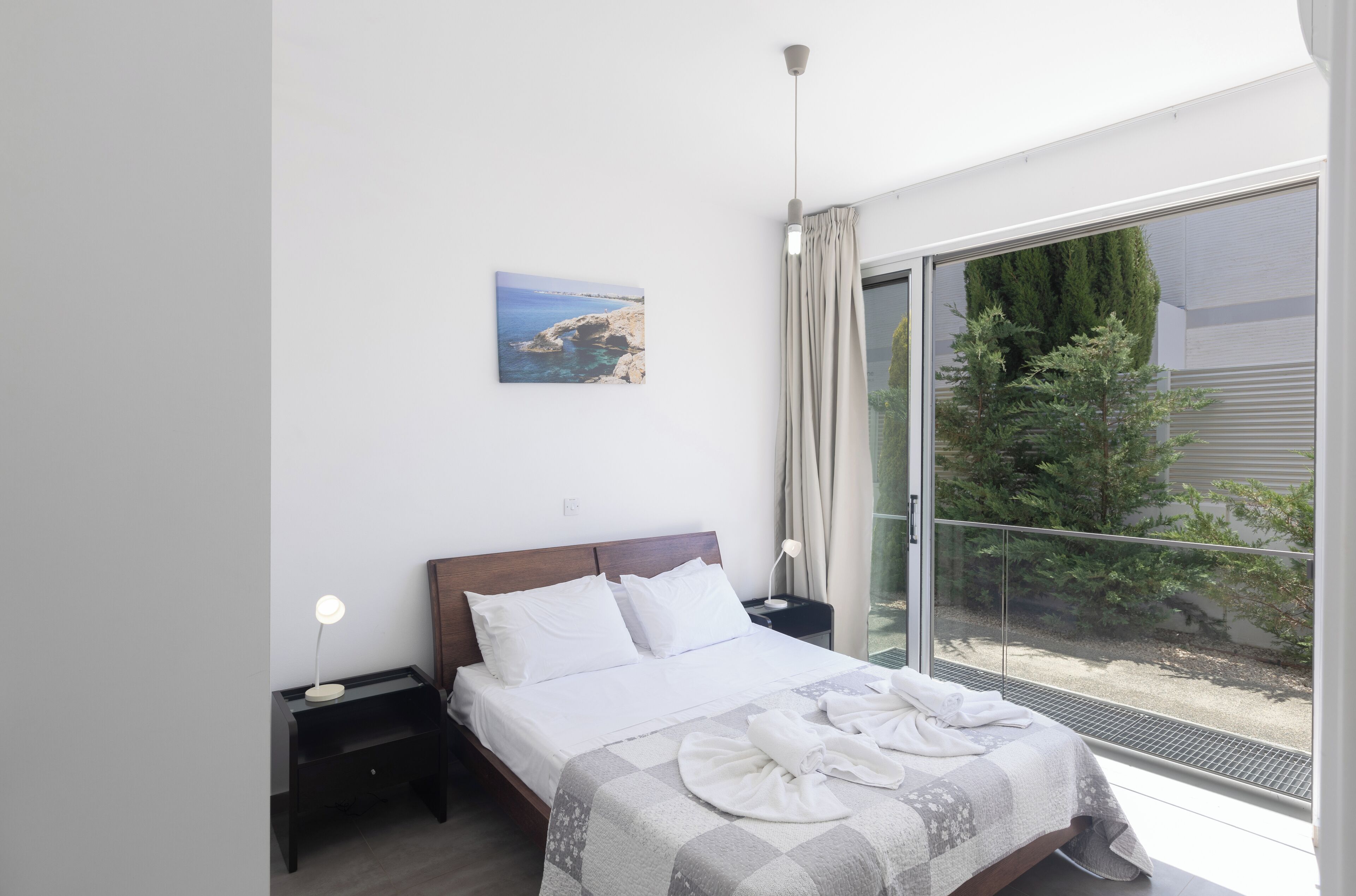 Een moderne slaapkamer met een groot bed, een nachtkastje, een bedlamp, een raam met uitzicht op groen en een schilderij aan de muur.